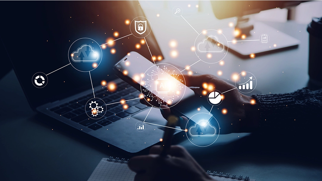 A hand holding a smartphone above a laptop, surrounded by digital icons symbolizing AI, data, and cloud technology.